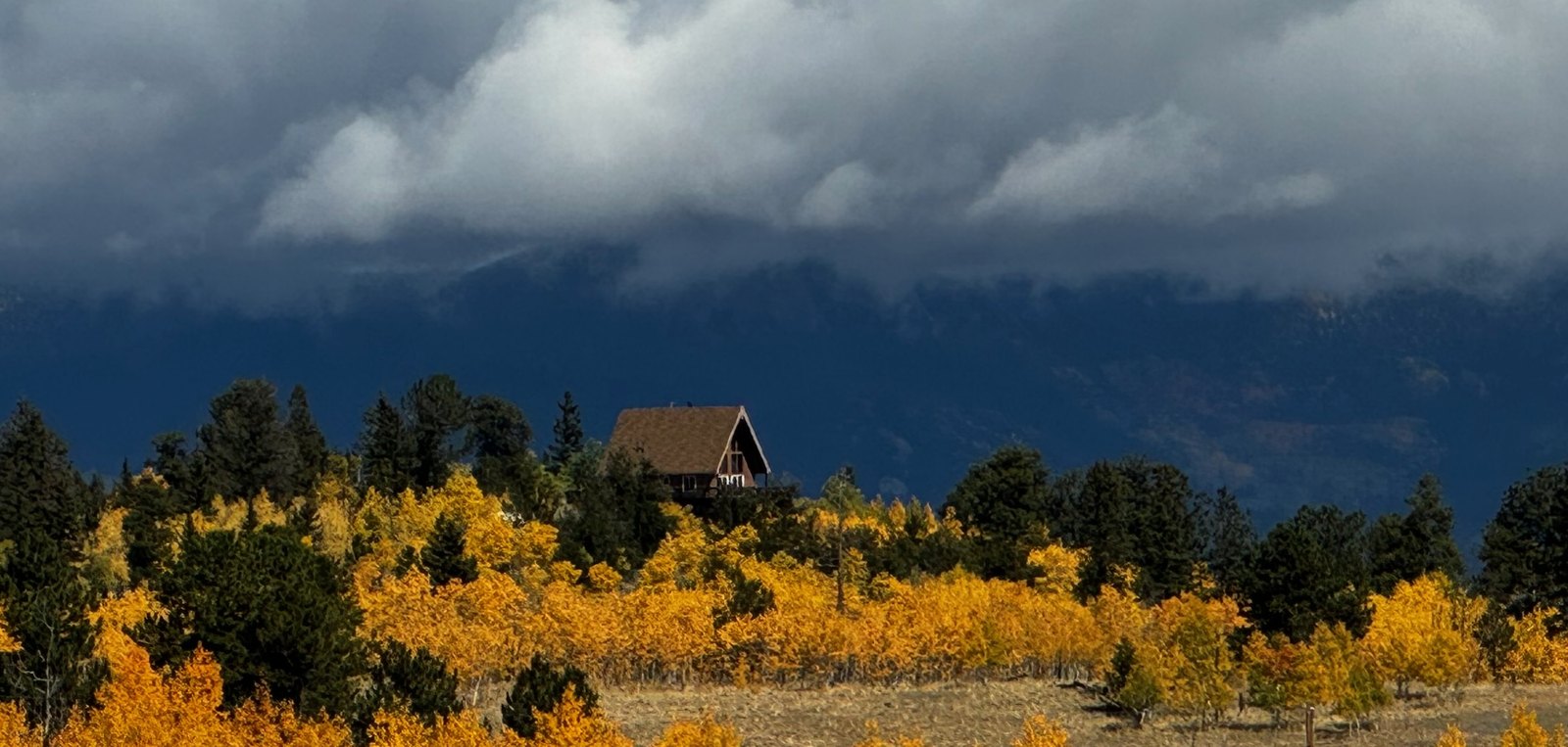 An A-frame cabin above golden aspens and dark pines, with storm clouds over the mountains behind