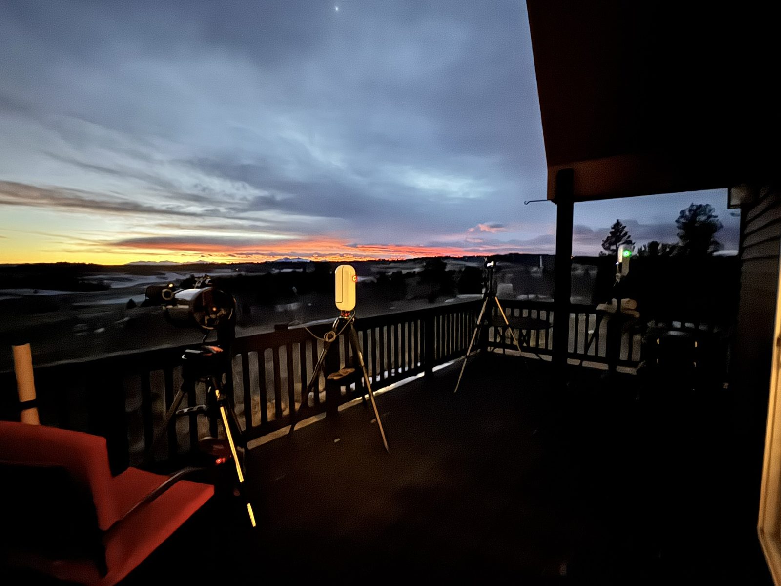 Multiple smart telescopes set up along a deck railing at dusk, a band of orange sunset over the Colorado hills behind them